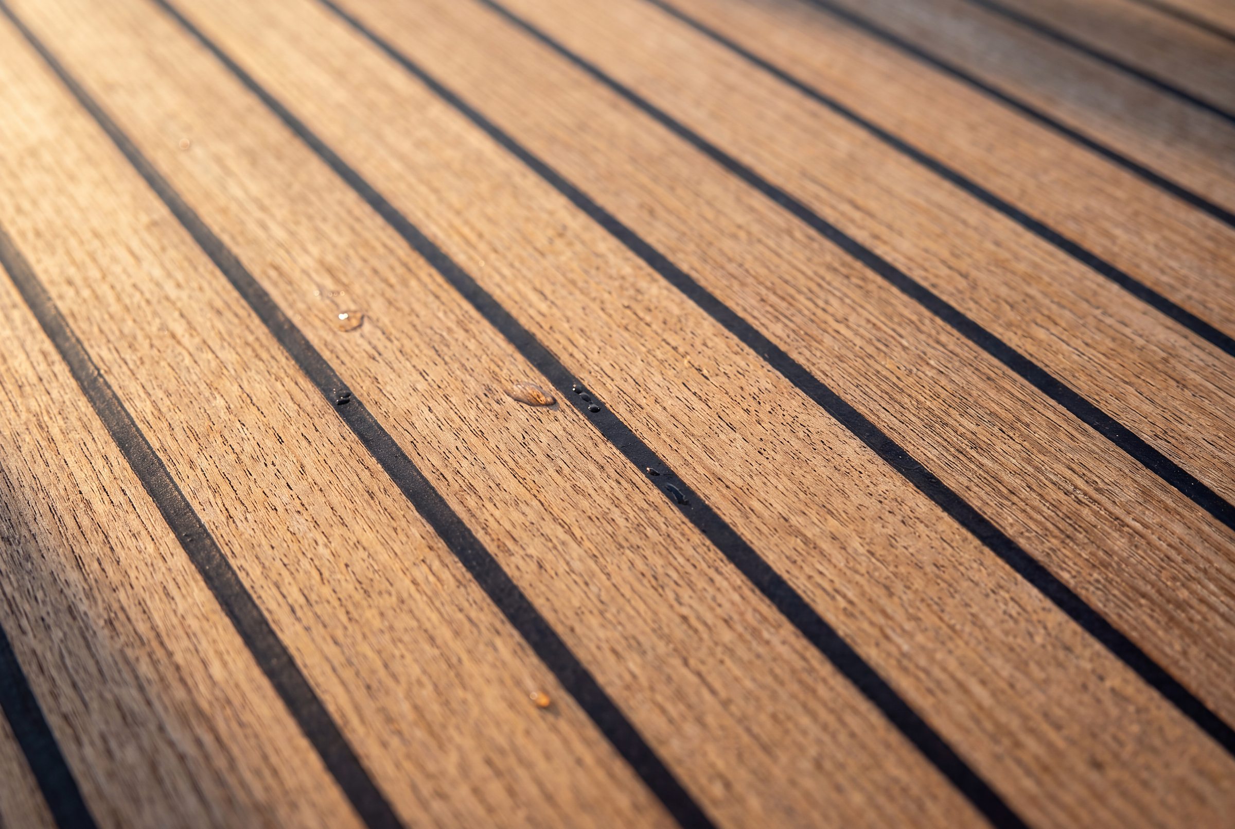 Visione Naviglia: macro di doghe di teak naturale dorato con fughe nere e poche gocce d'acqua