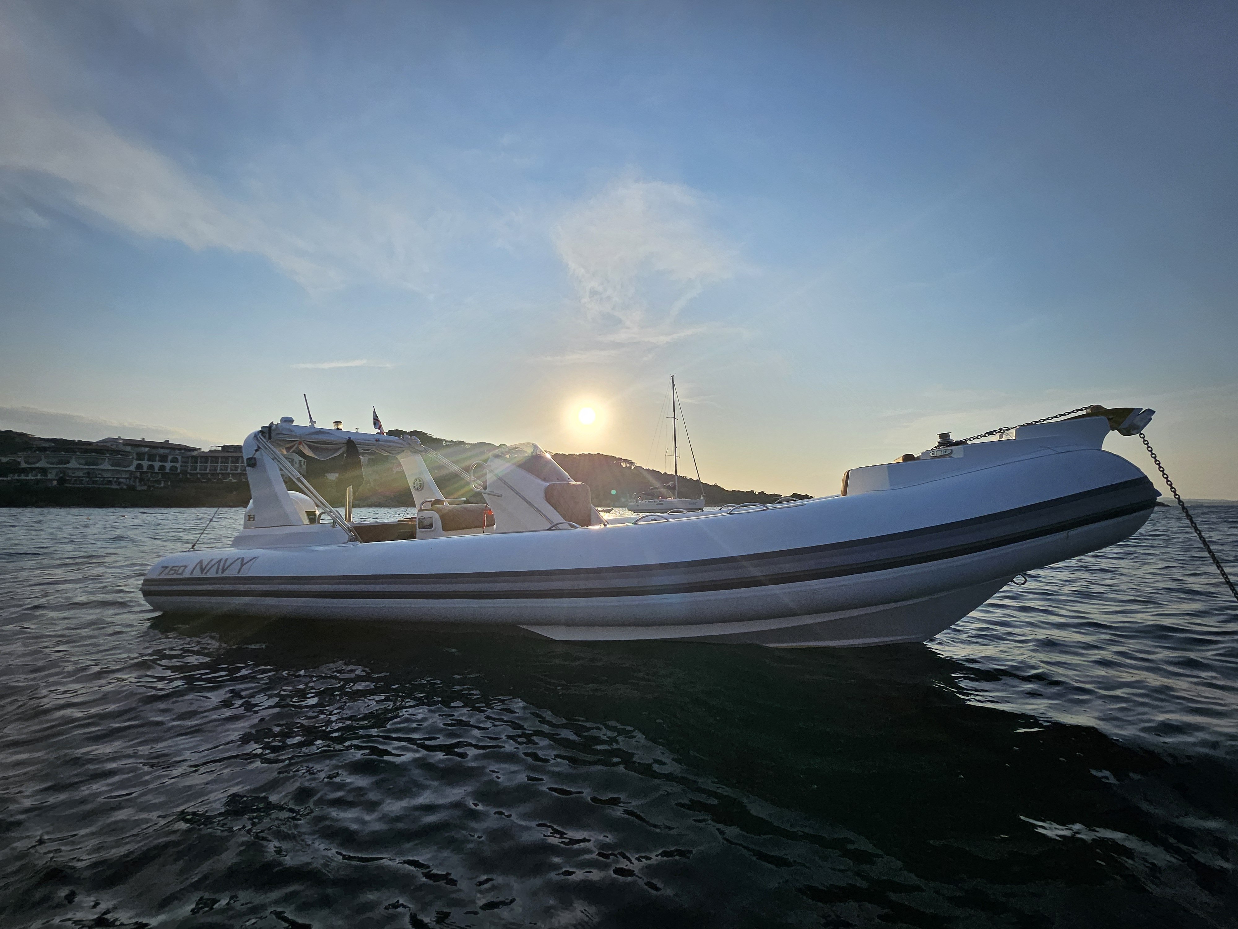 White NAVY 7.60 at anchor at sunset, low sun behind the roll bar and the anchor chain in the water