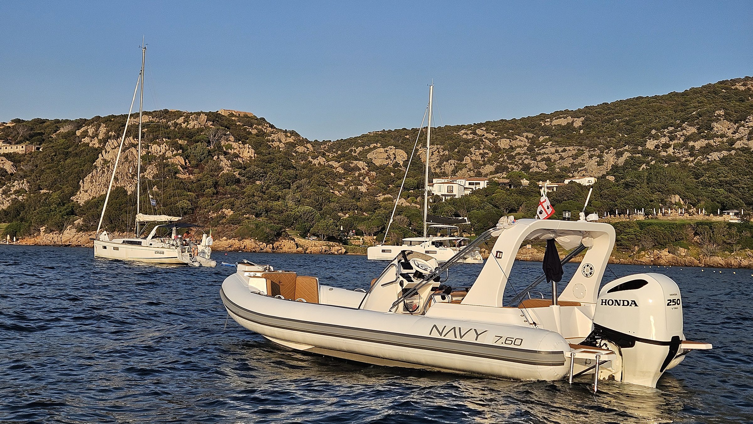NAVY 7.60 at anchor, stern three-quarter view, white Honda 250 and scrub-covered hills behind