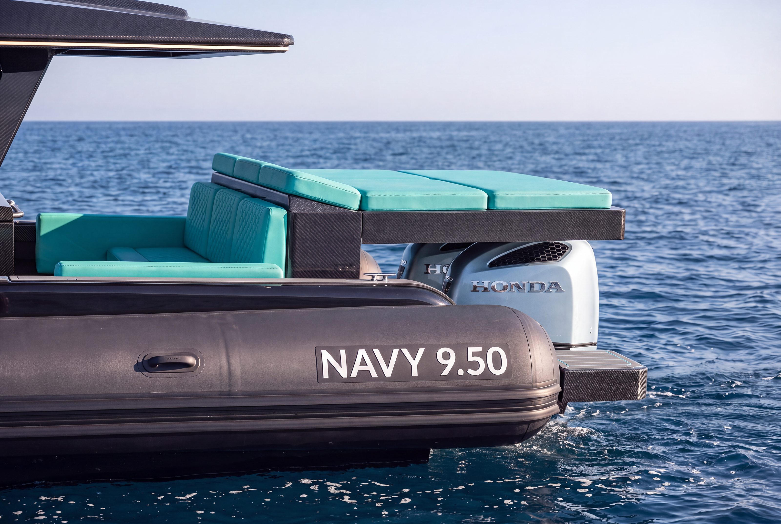Naviglia vision: the stern of the new NAVY 9.50 Cabin under way, with the NAVY 9.50 lettering on the black tube, the turquoise sofa and sunpad and, under the sunpad, the silver cowling of a Honda BF350